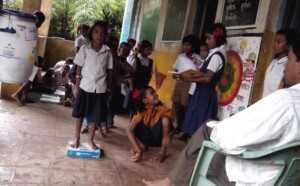 School kids undergo routine health record keeping. Girl on weighing scale and other girl maintaining records. 